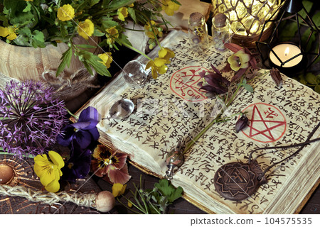 Still life with open magic book of spells, candle and medallion on witch ritual table. Occult, esoteric and witchcraft concept. Mystic background with vintage objects Still life with open magic book of spells, candle and medallion on witch ritual table. Occult, esoteric and witchcraft concept. Mystic background with vintage objects 104575535