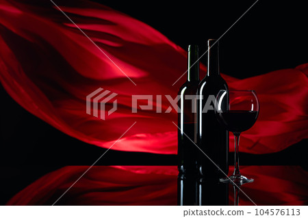 Wineglass and bottles of red wine on a black reflective background. Red satin curtain flutters in the wind. Wineglass and bottles of red wine on a black reflective background. Red satin curtain flutters in the wind. 104576113