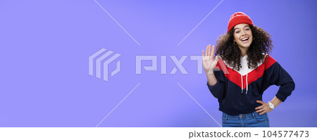 Hey nice to meet you. Charming female ski couch in cute red beanie with curly hair waving hello with palm and smiling broadly greeting newbies as teaching winter sports over blue background 104577473