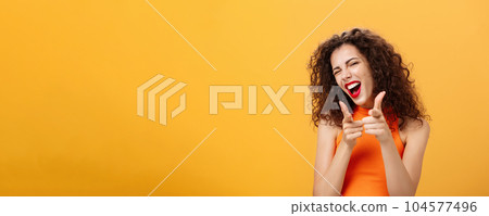 Hey what is up. Friendly and outgoing attractive curly-haired female with red lipstick pointing at camera with finger guns as if greeting or hinting friend winking at camera over orange background 104577496