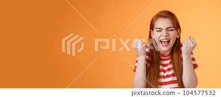 I did it. Charming lucky determined redhead female achieve success, fist pump celebrating, close eyes yell yes yeah, hooray gesture, triumphing victory, very good news, stand orange background 104577532