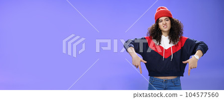 Snobbish and displeased stylish and cool street girl with curly hair in beanie and sweatshirt pointing down grimacing as feeling unsatisfied and disappointed posing against blue background 104577560