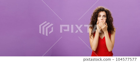 Cocnerned and shocked troubled adult woman with curly hairstyle in elegant evening red dress gasping from surprise and fear covering mouth with palms standing speechless over purple wall Cocnerned and shocked troubled adult woman with curly hairstyle in elegant evening red dress gasping from surprise and fear covering mouth with palms standing speechless over purple wall 104577577