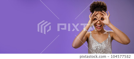 Playful funny good-looking african woman in afro hairstyle in silver glittering dress having fun show tongue smiling broadly make okay ok gesture on eyes look excited, standing blue background Playful funny good-looking african woman in afro hairstyle in silver glittering dress having fun show tongue smiling broadly make okay ok gesture on eyes look excited, standing blue background 104577582