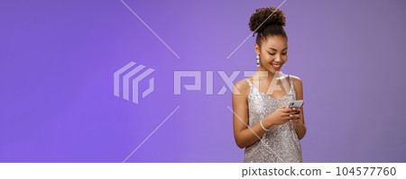 Charming elegant african-american woman in stylish glittering prom dress writing message calling friend hold smartphone look amused happy smile phone display using gadget app scroll party photos 104577760