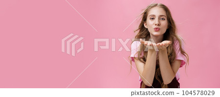 Portrait of sensual and tender gentle young wman with wavy natural hair bending towards camera facing wind holding palms near folded lips sending air kiss forward with flirty caring and loving look 104578029