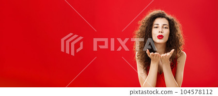 Woman rewards boyfriend with passionate wind kiss after romantic date. Portrait of sensual hot adult female in red elegant dress with curly hairstyle bending towards camera sending mwah flirty 104578112