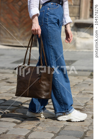 photo of a woman wearing blue jeans and white blouse with a brown leather handbag . 104578276