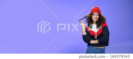 Stylish and snobbish arrogant curly-haired woman in warm beanie rolling curl on finger smirking and looking with contempt at camera, scorning person as being too cool over blue background Stylish and snobbish arrogant curly-haired woman in warm beanie rolling curl on finger smirking and looking with contempt at camera, scorning person as being too cool over blue background 104578343