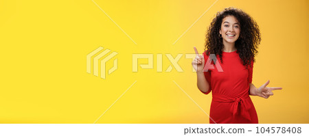 Charismatic cute and happy young curly-haired european female in red dress dancing and pointing up and right, showing sideways as presenting choices picking, making decision and smiling at camera 104578408