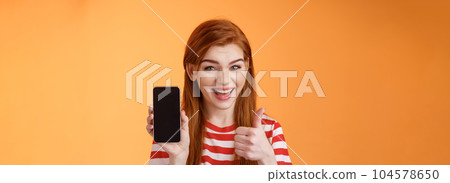 Cheerful assertive redhead woman recommend smartphone app, show phone display, thumb-up like approval sign, smiling broadly, suggest good online purchase, mobile advertisement 104578650