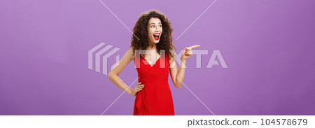Amused attractive european female with evening makeup in luxurious red dress and curly hairstyle laughing out loud entertained and happy pointing and gazing left intrigued over purple background Amused attractive european female with evening makeup in luxurious red dress and curly hairstyle laughing out loud entertained and happy pointing and gazing left intrigued over purple background 104578679