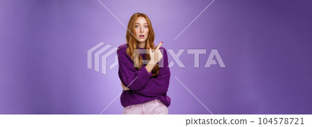 Astonished excited redhead woman open mouth and raise eyebrows questioned as seing shocking thing pointing at upper left corner, asking question impressed and surprised over purple wall 104578721