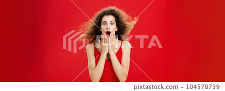 Attractive elegant young woman in red evening dress with curly hair flicking on wind saying wow being impressed and thrilled covering opened mouth from amazement standing over red background 104578739