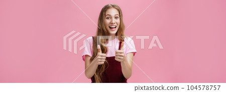 Friendly and outgoing pretty young woman in overalls with wavy natural hairstyle showing thumbs up gesture with cheer and joy smiling being supportive, liking idea, expressing positive opinion 104578757
