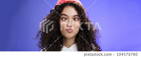 Headshot of funny and playful girl fooling around making faces not scared being funny folding lips like duck and squinting ice, aping standing childish and hilarious over blue wall in winter hat 104578760