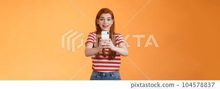 Amused excited redhead female record video thrilled see famous person, smiling amazed hold smartphone taking picture, tourist adore trip shooting everything during summer journey, orange background 104578837