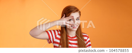Charismatic lovely redhead lucky female, smiling cute, peek camera tender coquettish gaze, hold palm face, look through fingers, stand orange background optimistic, cheerful mood 104578855