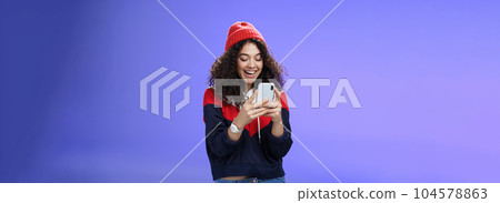 Joyful and sociable stylish caucasian woman with curls in warm beanie having fun laughing and chuckling as using smartphone typing funny message looking at mobile phone screen happily over blue wall 104578863