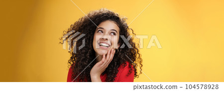 Happy positive pretty young woman with curly hair in red blouse laughing silly and carefree as gazing pleased at upper left corner, touching face, satisfied and delighted over yellow wall 104578928