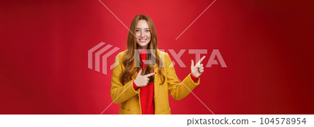 Cheerful friendly-looking and energized helpful redhead girl in yellow coat showing way pointing at upper left corner and smiling broadly with satisfied grin posing delighted over red background 104578954