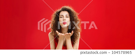 Woman rewards boyfriend with passionate wind kiss after romantic date. Portrait of sensual hot adult female in red elegant dress with curly hairstyle bending towards camera sending mwah flirty 104578965