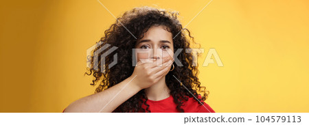 Close-up shot of concerned and insecure troubled woman keep silent scare of telling anyone her problem covering mouth with palm not to scream or slip word looking sad and sorrow over yellow background 104579113