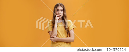 Girl worried mom react on new tattoo. Portrait of anxious and nervous silly insecure young female with wavy natural hairstyle biting finger panicking looking concerned and troubled over orange wall 104579224