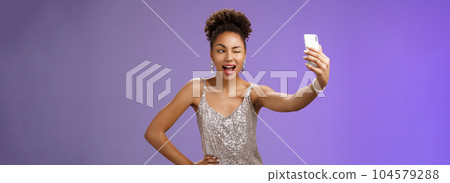 Stylish sassy african american b-day girl taking selfie new elegant silver shiny dress extend arm holding smartphone posing winking display screen amused having fun smiling broadly, blue background Stylish sassy african american b-day girl taking selfie new elegant silver shiny dress extend arm holding smartphone posing winking display screen amused having fun smiling broadly, blue background 104579288