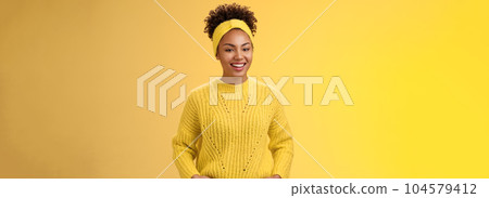 Friendly outgoing relaxed young african-american beautiful girl headband forehead in sweater hold hands pockets smiling broadly have fun talking interesting communication, standing yellow wall 104579412