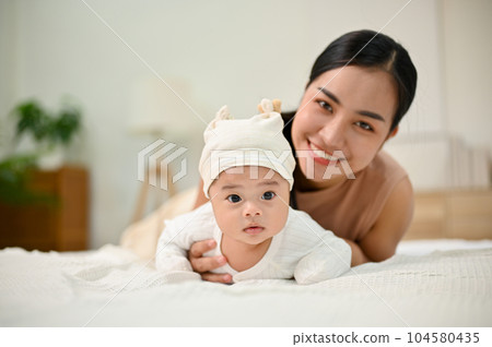Sweet family moment. Young Asian mom lay down with her baby, smiling to camera. 104580435