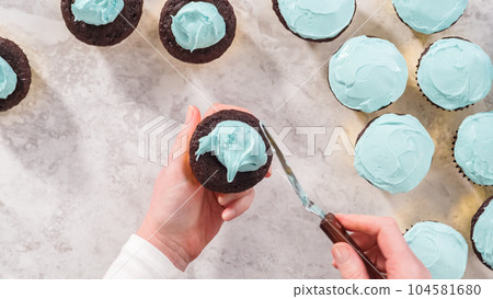 Flat lay. Step by step. Scooping blue buttercream frosting on top of the chocolate cupcakes. 104581680