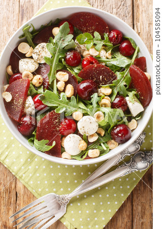 Fresh summer salad of arugula, baked beets, cherries, mozzarella, hazelnuts close-up in a bowl. Vertical top view 104585094