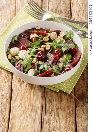 Dietary vitamin salad of arugula, baked beets, cherries, mozzarella, hazelnuts close-up in a bowl. Vertical 104585101