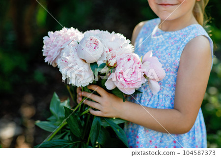 Cute adorable little preschool girl with huge bouquet of blossoming pink peony flowers. Portrait of smiling preschool child in domestic garden on warm spring or summer day. Summertime. 104587373
