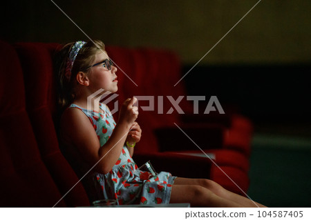 Little preschool girl with glasses watching cartoon movie or film in cinema. Happy excited child. Family activity with children. Little preschool girl with glasses watching cartoon movie or film in cinema. Happy excited child. Family activity with children. 104587405