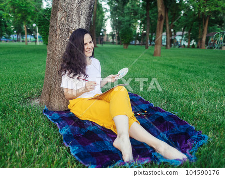 Art therapy. Woman draws in the park. Girl sits with her back against a tree and looks into distance. Restoration of nervous system with help of drawing. Concept of relaxation and meditative state 104590176