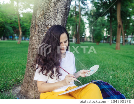 Art therapy. Woman draws in the park. Girl sits with her back against a tree and looks into distance. Restoration of nervous system with help of drawing. Concept of relaxation and meditative state 104590178