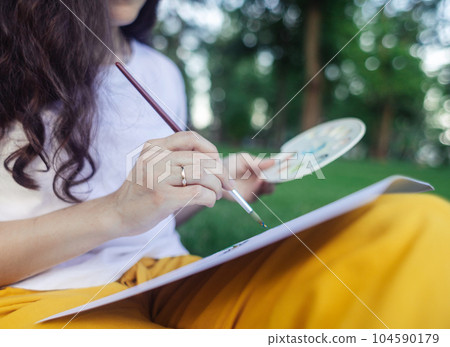Art therapy. Woman draws in the park. Girl sits with her back against a tree and looks into distance. Restoration of nervous system with help of drawing. Concept of relaxation and meditative state 104590179