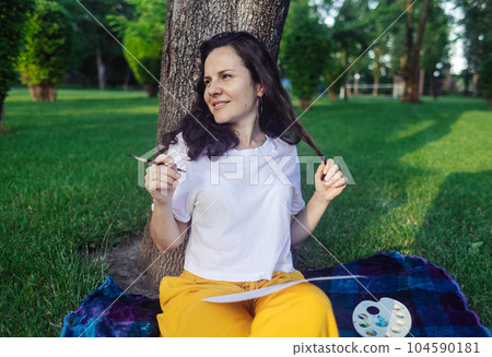 Art therapy. Woman draws in the park. Girl sits with her back against a tree and looks into distance. Restoration of nervous system with help of drawing. Concept of relaxation and meditative state Art therapy. Woman draws in the park. Girl sits with her back against a tree and looks into distance. Restoration of nervous system with help of drawing. Concept of relaxation and meditative state 104590181