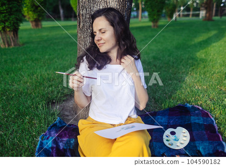 Art therapy. Woman draws in the park. Girl sits with her back against a tree and looks into distance. Restoration of nervous system with help of drawing. Concept of relaxation and meditative state 104590182