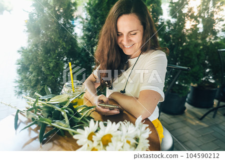 Happy smiling girl sits at a table in a cafe after a romantic date. There is a bouquet of daisies on table. Woman in a brown hat looks at her phone, reads correspondence and remembers happy moments Happy smiling girl sits at a table in a cafe after a romantic date. There is a bouquet of daisies on table. Woman in a brown hat looks at her phone, reads correspondence and remembers happy moments 104590212