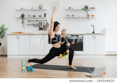Lady doing walking lunges with kid on knee in kitchen 104594920