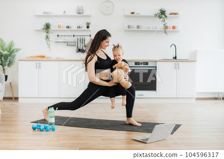 Mother doing walking lunges with baby and teddy bear indoors 104596317