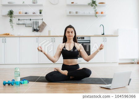 Woman doing yoga to music via laptop in kitchen interior Woman doing yoga to music via laptop in kitchen interior 104596366