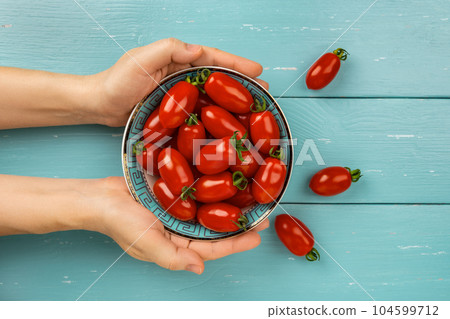 Woman hands hold bowl of red small tomatoes over turquoise wood background. Woman hands hold bowl of red small tomatoes over turquoise wood background. 104599712