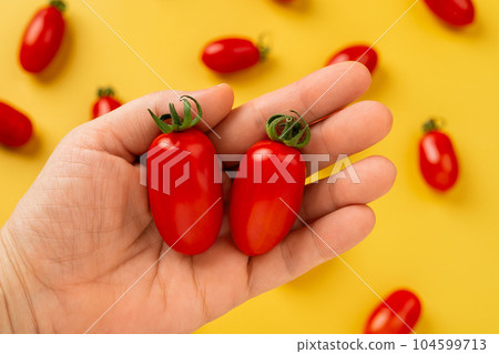 Female hand holds two red Ornela cherry tomatoes over yellow background. 104599713