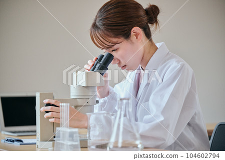 A young woman in a white coat looking through the lens of a microscope and observing A young woman in a white coat looking through the lens of a microscope and observing 104602794