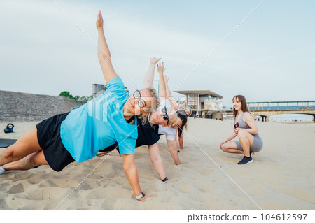 Women of various ages doing fitness workouts in class exercise with coach on the city beach on sunset. Ladies doing a side plank. Sport for health and wellbeing. Active lifestyle. Selective focus 104612597