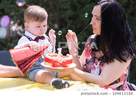 blissful toddler boy with mother celebrate his birthday, with birthday hat and a cake and on candle. outdoor blissful toddler boy with mother celebrate his birthday, with birthday hat and a cake and on candle. outdoor 104613188